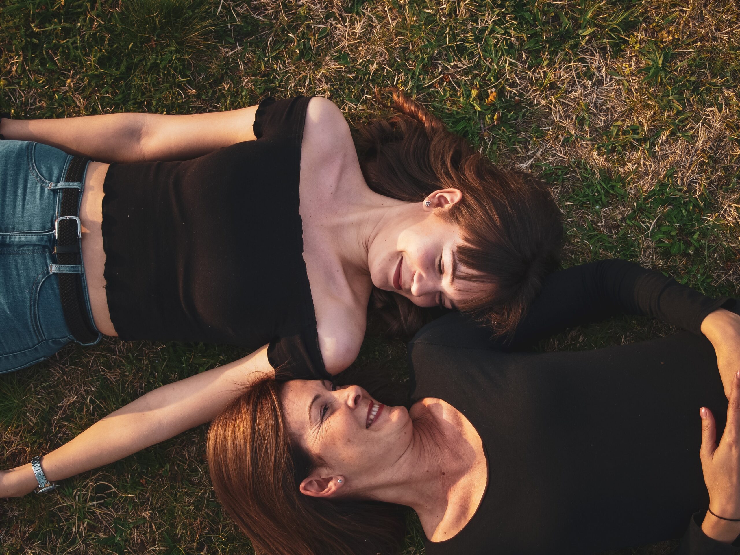 2 vrouwen die in het gras liggen, zon op de huid. Ze kijken elkaar aan terwijl ze lachen.